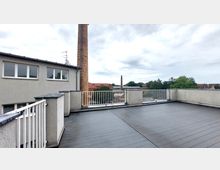 Flachdach-Terrasse mit dunkelgrauem Bodenbelag, umgeben von niedrigen Betonbrüstungen mit weißen Metallgeländern; im Hintergrund eine Ziegelschornstein-Struktur, Wohnhäuser mit roten Dächern und grüne Bäume unter wolkigem Himmel.