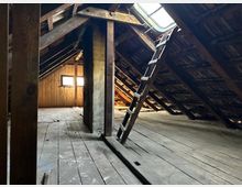 Alter Dachboden mit sichtbaren Holzbalken, Holzfußboden und zwei kleinen Fenstern an einer holzverkleideten Wand. Eine einfache Holzleiter führt zu einem Dachfenster, durch das Tageslicht einfällt.