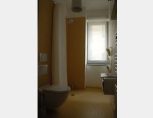 Kleines Badezimmer mit beige-gelben Fliesen, einer Wandtoilette, einer Duschkabine mit weißem Duschvorhang und einem Fenster mit blickdichtem Rollo. Ein Handtuchheizkörper und ein kleines Waschbecken mit Ablagefläche sind ebenfalls vorhanden.
