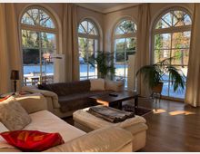 Wohnzimmer mit großen, halbkreisförmigen Fenstern, die viel Tageslicht hereinlassen und auf einen verschneiten Garten blicken. Das Zimmer ist mit beigen Sofas, einem Holztisch, Pflanzen und Vorhängen gestaltet, während der Holzboden eine warme Atmosphäre schafft.