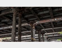 Unterseite einer alten Eisenbahnbrücke mit verzierten gusseisernen Säulen. Die Struktur zeigt Spuren von Rost und Verwitterung, mit stählernen Verstrebungen und Nieten, die die Konstruktion stützen.