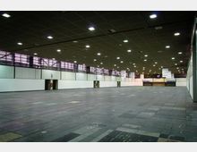 Geräumige, leere Messehalle mit hoher Decke, rechteckigem Grundriss und grauem Bodenbelag aus quadratischen Platten; Wände sind hell und mit Türen sowie großen Fenstern im oberen Bereich gestaltet, die schwaches Tageslicht durchlassen. Deckenleuchten sorgen für helle künstliche Beleuchtung, im Hintergrund hängt ein Schild mit der Nummer „11“.