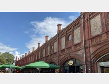 Backsteingebäude mit reich verzierten Fassadenelementen und ovalen Fenstern, vermutlich Teil eines historischen Bahnhofs oder Markthallenkomplexes, im Vordergrund mehrere grüne Sonnenschirme und geschäftiger Außenbereich.