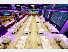 Großer Veranstaltungsraum mit mehreren langen, weiß gedeckten Tischen und hellen Polsterstühlen, arrangiert in symmetrischen Reihen. Im Hintergrund befinden sich eine Bar und große Fensterfronten, die den Raum mit Tageslicht beleuchten.