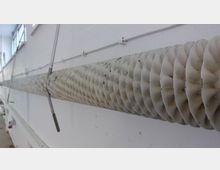 Nahaufnahme einer langen, weißen Heizungs- oder Lüftungsröhre mit einem wellenförmigen Design, die an einer hellen Wand mit sichtbaren Kabeln und Halterungen montiert ist. Im Hintergrund sind Fenster und eine industrielle Umgebung erkennbar.