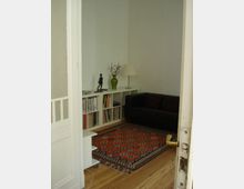 Ein kleiner Wohnraum mit Holzboden, einem dunklen Ledersofa, einem weißen Regal voller Bücher und Schallplatten sowie einer Tischlampe auf dem Regal. Ein gemusterter Teppich bedeckt den Boden, und eine geöffnete Tür mit einem goldfarbenen Griff führt in den Raum.