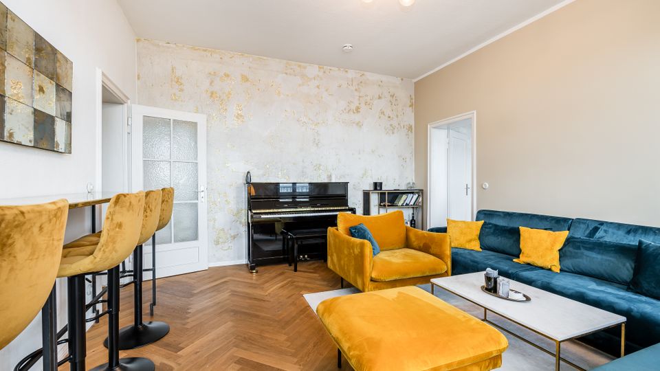 Modern eingerichtetes Wohnzimmer mit Fischgrät-Holzboden, samtigem Ecksofa in Petrol und gelben Sesseln samt Hocker um einen rechteckigen Couchtisch. An der Rückwand steht ein schwarzes Klavier vor einer hell gemusterten Akzentwand; links befindet sich ein schmaler Bartisch mit hohen Stühlen und zwei Türen führen in angrenzende Räume.