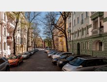 Pflastersteinstraße in einem Wohngebiet mit mehrstöckigen Altbauten in verschiedenen Farben, darunter Weiß, Gelb und Grün, mit dekorativen Fassaden. Bäume säumen die Straße, und Autos parken auf beiden Seiten. Im Hintergrund ist ein rotes Backsteingebäude erkennbar.