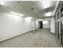 Ehemaliger leerstehender Verkaufsraum mit weißen Wänden, grauem Fliesenboden und einer Rasterdecke mit integrierter Leuchtstoffbeleuchtung. Auf der rechten Seite befinden sich großflächige Glasfenster oder Türen.