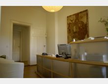 Ein schlicht eingerichteter Büro- oder Empfangsraum mit einem Schreibtisch aus Holz und Milchglas, darauf ein Computer und ein Telefon. Im Hintergrund befinden sich weiße Doppeltüren sowie ein abstraktes Kunstwerk an der Wand; rechts steht eine grüne Pflanze.