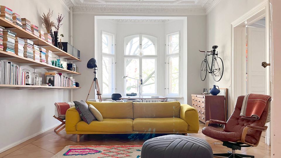 Bright living room with ornate ceiling moldings and a large bay window, featuring a mustard-yellow sofa, patterned rug, and a leather lounge chair. Wall-mounted bookshelves line one side, and a bicycle hangs above a wooden chest near a doorway.