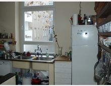 Kleine Küche mit einem Fenster, das zu einem Wohnhaus blickt. Zu sehen sind eine Spüle mit Arbeitsfläche, ein Kühlschrank, Regale mit Geschirr sowie diverse Küchenutensilien.