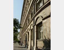 Fassade eines historischen Gebäudes mit hellem Stein und Rundbogenarchitektur, entlang einer Baum gesäumten Straße mit Gehweg und parkendem Motorrad.