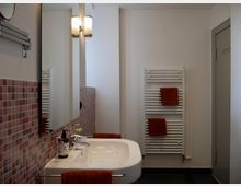 Modernes Badezimmer mit einer weißen Wand, einem Waschbecken mit Spiegel, rosafarbenen Mosaikfliesen und einem weißen Heizkörper, an dem orangefarbene Handtücher hängen. Links befindet sich eine Wandbeleuchtung, rechts eine geschlossene Tür.