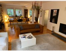 Wohnzimmer mit Holzfußboden, einer braunen Ledercouch und einem weißen Couchtisch in der Vordergrundansicht. Im Hintergrund befinden sich ein Esstisch mit Stühlen, eine große Fensterfront mit Vorhängen, ein Bücherregal, sowie ein Kronleuchter und eine weiße Wand mit Kamin.