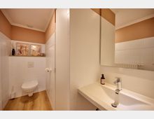 Modernes Gäste-WC mit beigefarbener Wand und weiß gefliesten Oberflächen. Zu sehen sind eine Hängetoilette mit Holzoptik-Boden, ein kleines Waschbecken mit Spiegel sowie Dekorationen wie Bilder und ein Duftspender auf einem Wandregal.