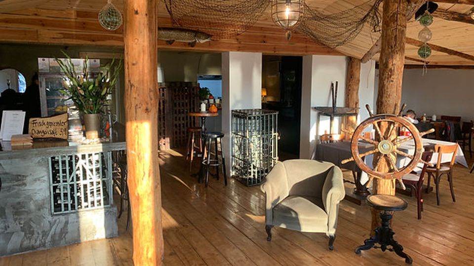 Gemütlicher Innenraum eines Cafés oder Restaurants mit maritimem Dekor, darunter Holzbalken, ein Schiffssteuerrad, Fischernetze an der Decke und Holzboden. Ein Tresen aus Stein und Holzmöbel ergänzen das rustikale Ambiente.
