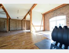 Geräumiger Innenraum mit Holzbalken und Backsteinwänden, kombiniert mit hellen weißen Wänden und einem großen Fensterbogen, der Tageslicht hereinlässt. Der Boden ist aus poliertem Holz, und im Vordergrund steht ein modernes, schwarzes Ofenmodell.