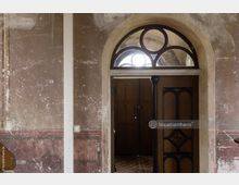 Verwitterter Innenraum mit einer hölzernen Tür im Mittelpunkt, die ein geometrisch verziertes Oberlicht und einen abgenutzten Türrahmen mit schwarzen Metallakzenten aufweist. Die Wände sind grob verputzt und zeigen Spuren von Abblätterungen, während der Boden aus Ziegelsteinen besteht.