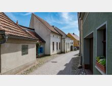 Pflastersteingasse mit kleinen Wohnhäusern in einem historischen Viertel. Die Häuser haben helle Fassaden, teils mit bunten Fensterläden, und rote Ziegeldächer; rechts sind Blumenkästen an einem Gebäude angebracht.