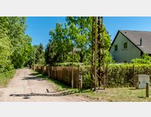 Eine ruhige, unbefestigte Sandstraße, gesäumt von Bäumen und einem hölzernen Zaun, führt an einem Wohnhaus mit grauer Fassade und Schrägdach vorbei. Im Vordergrund stehen ein Straßenschild mit der Aufschrift "Mozartallee" und ein Strommast aus Metall.