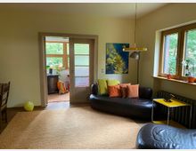 Wohnzimmer mit cremefarbenen Wänden, einem großen Teppich und einer schwarzen Ledercouch mit bunten Kissen. Im Hintergrund führt eine Glastür zu einem angrenzenden Raum mit großen Fenstern, und rechts sind weitere Fenster mit Blick ins Grüne.