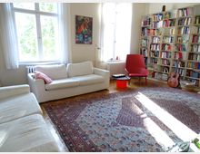 Helles Wohnzimmer mit großen Fenstern, weißen Sofas und einem roten Sessel. Im Hintergrund steht ein Bücherregal, daneben eine Akustikgitarre; am Boden liegt ein großer, gemusterter Teppich.