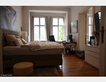 Schlafzimmer mit einem großen Bett auf der linken Seite, Parkettboden und hohen Decken. Im Raum stehen ein Schreibtisch mit Computer am Fenster und eine Kommode, während Tageslicht durch große Fenster mit weißen Vorhängen einfällt.