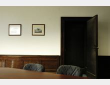 Ein Besprechungsraum mit einer Holzvertäfelung an der Wand, einem großen Holztisch und gepolsterten Stühlen. An der weiß gestrichenen Wand hängen zwei gerahmte Bilder, rechts ist eine halb geöffnete Holztür zu sehen.