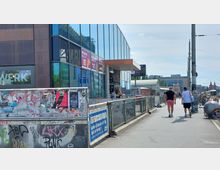 Moderne Fußgängerpassage entlang eines Gebäudes mit einer Fassade aus Glas und braunem Metall, auf der Werbeschilder angebracht sind. Im Vordergrund eine Brüstung mit Graffiti und Plakaten, im Hintergrund Fahrräder, Straßenbahnkabel und Stadtgebäude.