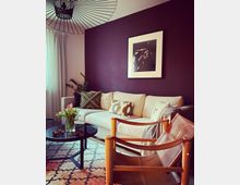 Wohnzimmer mit einer cremefarbenen Eckcouch, dekoriert mit gemusterten Kissen vor einer dunkelvioletten Wand, an der ein gerahmtes Bild hängt. Im Vordergrund ein runder Glastisch mit Blumen, ein Holzstuhl mit Ledersitz und ein gemusterter Teppich; modernes Hängelicht an der Decke.