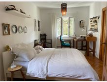 Schlafzimmer mit Doppelbett, weißer Bettwäsche und einer weißen Wand, dekoriert mit Bildern und Metallplatten. Im Hintergrund ein Schreibtisch mit einem Stuhl vor einem Fenster, das Tageslicht hereinlässt, sowie ein hölzerner Fußboden und eine hängende Lampe mit Perlen.