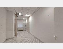 Ein leerer, rechteckiger Raum mit weißen Wänden und einem Boden aus kleinen weißen Fliesen. Der Raum hat eine abgehängte Decke mit eingebauten Leuchtpaneelen, ein Fenster mit weißem Lamellenvorhang und eine Heizung darunter.