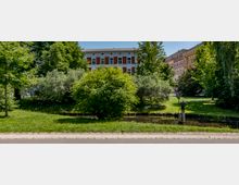 Ein kleiner Park mit einer Rasenfläche, Bäumen und Sträuchern. Im Vordergrund verläuft ein schmaler Wassergraben, daneben steht eine kleine Statue auf einem Sockel. Im Hintergrund sind mehrstöckige Gebäude mit weißen und roten Fassaden zu sehen.