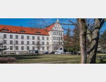 Historisches Gebäude mit weißer Fassade, zahlreichen Fenstern und einem roten Satteldach, umgeben von einer Rasenfläche mit Bäumen. Im Vordergrund sind eine Baumgruppe sowie parkende Autos entlang einer Straße sichtbar.