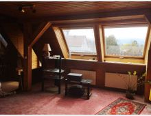 Ein Dachgeschosszimmer mit holzverkleideten Wänden und Decke, ausgestattet mit großen schrägen Dachfenstern, die viel Tageslicht hereinlassen. Im Raum befinden sich ein Regal, eine Tischlampe, Heizkörper unter den Fenstern und ein Teppich auf einem rosa Teppichboden.