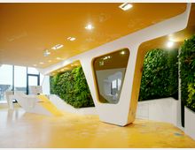 Modernes Büro mit gelb-orangefarbenem Boden und Decke, wandhohen vertikalen Pflanzenwänden und futuristischer weißer Rezeption in geschwungenem Design. Große Fenster lassen Tageslicht herein und bieten Blick nach draußen.