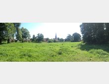 Grüne Wiese mit einzelnen Bäumen und Häusern im Hintergrund; eine Kirche mit einem hohen, spitzen Kirchturm ragt zentral hervor. Die Szene ist von Bäumen umgeben, und der Himmel ist klar und sonnig.