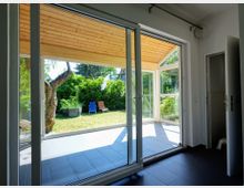 Blick aus einem Innenraum mit dunklem Fliesenboden durch eine große Glastür auf eine überdachte Terrasse mit Holzdecke und einen Garten mit Rasen, Bäumen und Gartenstühlen. Im Hintergrund sind grüne Hecken und Pflanzen erkennbar.