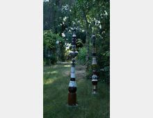 Zwei kunstvolle Skulpturen aus gestapeltem Geschirr und Keramik stehen auf einer grasbewachsenen Fläche in einem Garten. Der Garten ist von Bäumen und Büschen umgeben, die eine schattige, naturnahe Umgebung schaffen.