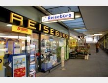 Außenansicht eines Reisebüros mit gelbem Leuchtschriftzug „Reisebüro“ und Glasfassade, die mit Reiseprospekten, Postern und Werbematerialien dekoriert ist. Daneben befinden sich ein Blumenstand und Werbeschilder für „Lotto“.