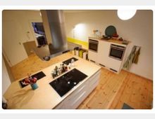 Moderne Küche mit hellen Holzböden, einer zentralen Kochinsel mit eingebautem Herd und Dunstabzugshaube. Im Hintergrund sind zwei integrierte Backöfen in einer weißen Wandverkleidung sowie eine gelbe Rückwand und offene Ablageflächen zu sehen.