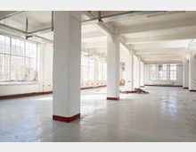 Helle, leere Industriehalle mit hohen Decken, weißen Stützpfeilern und freiliegenden Kabeltrassen. Große Sprossenfenster entlang der Außenwände lassen viel Tageslicht auf den Betonboden fallen; teilweise sind Baustellenabsperrbänder und Schutt zu sehen.