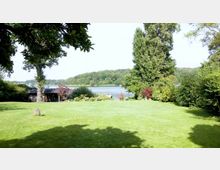 Grüne Wiese mit vereinzelten Sträuchern und Bäumen im Vordergrund, dahinter ein kleiner Holzschuppen sowie ein See, der von dicht bewaldeten Hügeln am Horizont umgeben ist.