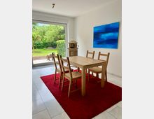 Heller Essbereich mit Holztisch und vier Holzstühlen auf einem roten Teppich, weißen Wänden und Fliesenboden. Große Schiebetür öffnet den Blick in einen grünen Garten; an der Wand hängt ein blaues abstraktes Bild, daneben steht ein antiker Ofen.