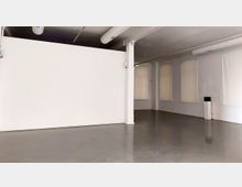 Minimalistischer Innenraum mit glatten Betonboden und weißen Wänden. Der Raum verfügt über hohe Fenster mit geschlossenen weißen Rollos, sichtbare Lüftungsrohre an der Decke und einen schlichten rechteckigen Pfeiler.