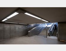 Unterirdischer U-Bahn-Eingang mit Betonboden und grauen Metallwänden. Im Hintergrund führt eine Treppe sowie eine Rolltreppe nach oben, beide von neonbeleuchteten Deckenlampen erhellt; eine Person steht auf der Rolltreppe.