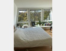 Ein Schlafzimmer mit einem großen Bett, das zentral positioniert ist. Im Hintergrund befinden sich drei große Fenster mit Blick auf eine Baumlandschaft, darunter eine Fensterbank mit Pflanzen, Büchern und Dekorationsgegenständen.