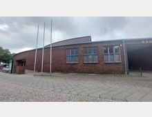 Backstein-Gebäude mit gebogener Fassade, mehreren großen Fenstern mit blau-roten Rahmen und daneben drei leeren Fahnenmasten. Im Vordergrund befindet sich ein gepflasterter Gehweg, im Hintergrund sind Bäume erkennbar.