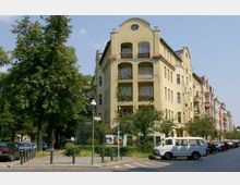 Straßenszene mit einem mehrstöckigen, gelben Wohngebäude im späten Jugendstil, das Balkone mit Pflanzen und Rundbogenfenster aufweist. Bäume und parkende Autos säumen die gepflasterte Straße, Straßenschilder und eine klassische Straßenlaterne stehen an einer Kreuzung.